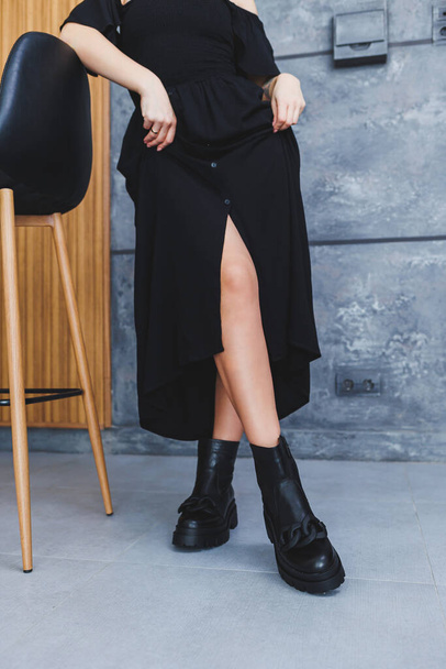 Schwarze Lederstiefel ohne Absatz mit Schnalle, schlanke Frauenbeine und ein Holzstuhl auf grauem Hintergrund. Das Konzept einer neuen Kollektion von Schuhen für den Frühling. Frau im schwarzen Kleid mit Schlitz - Foto, Bild