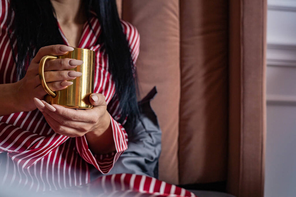 Closeup beauty female hands in pajamas holding golden metallic mug with hot beverage relaxing on bed - Foto, Imagen