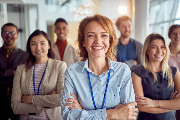 Close-up shot of cheerful female manager and her young team standing in a pleasant atmosphere in the company building hallway and posing for a photo. Business, people, company - Фото, изображение