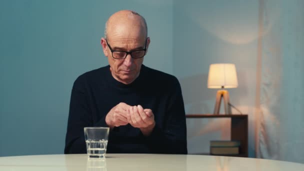 Homme âgé avec des lunettes assis à la table à la maison mange une pilule cardiaque - Séquence, vidéo