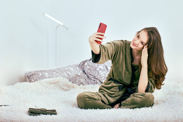 Online call with smartphone by young woman in dressing gown who sits on bed. - Фото, зображення