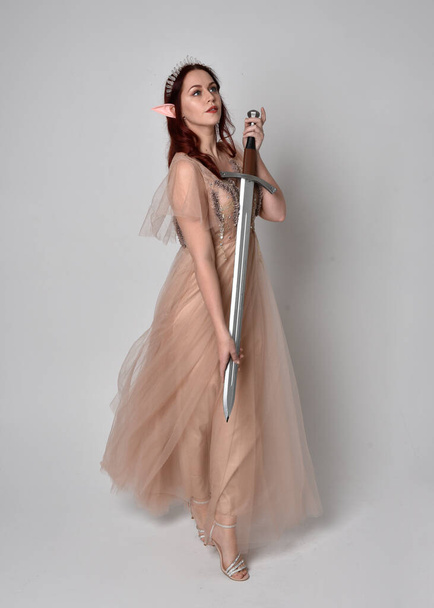  Full length portrait of pretty female model with red hair wearing glamorous fantasy tulle gown, crown. Holding a sword weapon, on studio background. - Photo, Image