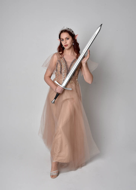  Full length portrait of pretty female model with red hair wearing glamorous fantasy tulle gown, crown. Holding a sword weapon, on studio background. - Photo, Image