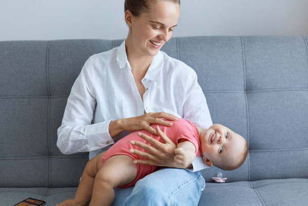 Indoor shot of smiling happy woman wearing white shirt and jeans sitting on sofa with daughter and expressing happy emotions, spending time with child during maternity leave. - Photo, image
