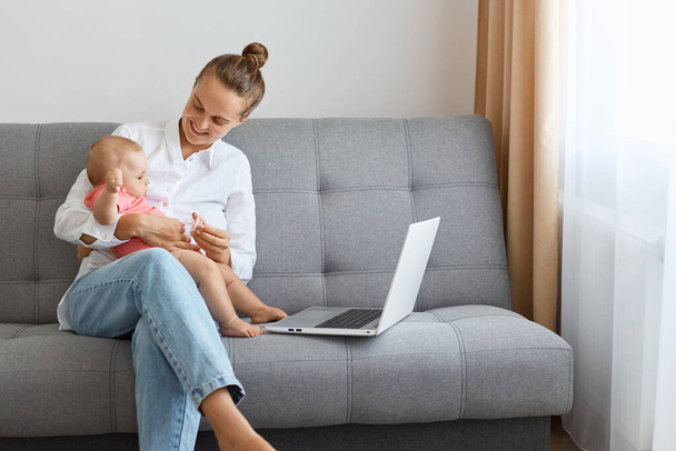 Portrait of smiling positive young mother with baby, woman with bun hairstyle wearing white shirt and jeans playing with toddler kid and working on computer. - 写真・画像