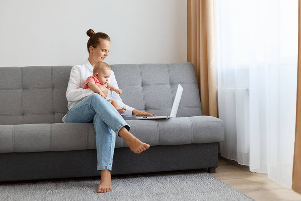 Full length portrait of slim Caucasian woman with bun hairstyle wearing white shirt and jeans working on notebook and holding infant child on her legs, watching video or checking social networks. - Foto, immagini