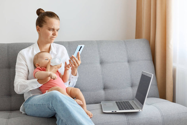 Horizontal shot of serious woman with bun hairstyle wearing white shirt and jeans, posing with her baby daughter and using phone, working on laptop, talking with business partner. - Fotó, kép