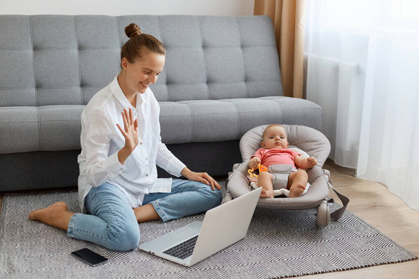 Horizontal shot of delighted woman with bun hairstyle wearing white shirt and jeans, sitting on floor with hey newborn baby and having video call on laptop, waving hand, saying hello. - Foto, afbeelding