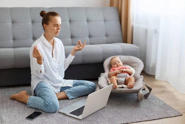 Indoor shot of attractive tired woman with bun hairstyle wearing white shirt and jeans, sitting on floor in front of laptop and looking after baby, doing yoga to calm down. - Valokuva, kuva