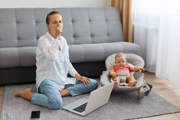 Image of tired sleepy woman with bun hairstyle wearing white shirt and jeans, female freelancer working online on laptop and looking after daughter, yawning. - Photo, image
