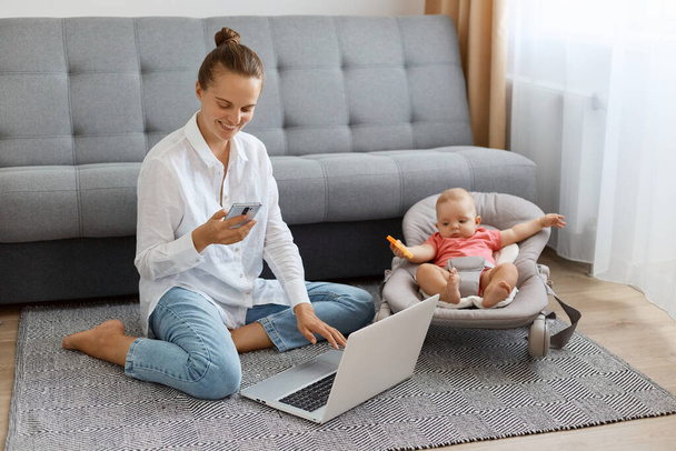 Full length portrait of satisfied woman with bun hairstyle wearing white shirt and jeans, young mother looking after baby, using smart phone and personal computer. - Fotoğraf, Görsel