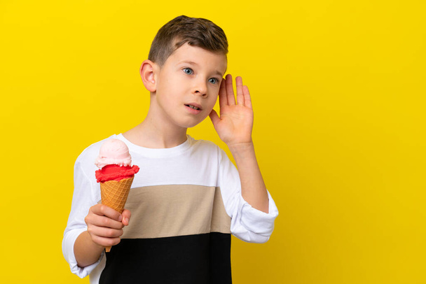 Petit garçon caucasien avec une crème glacée cornet isolé sur fond jaune écoutant quelque chose en mettant la main sur l'oreille - Photo, image