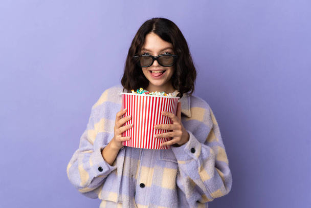 Teenager Ukrainian girl isolated on purple background with 3d glasses and holding a big bucket of popcorns - Foto, afbeelding