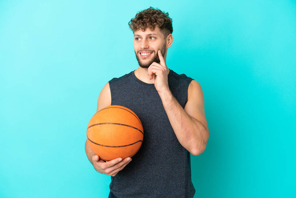 Hezký mladý muž hraje basketbal izolované na modrém pozadí myšlení nápad při pohledu nahoru - Fotografie, Obrázek