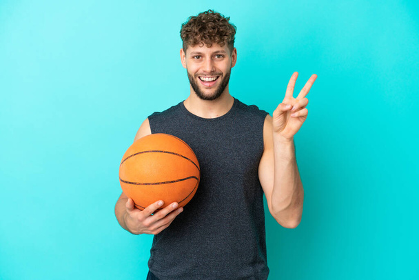 Beau jeune homme jouant au basket isolé sur fond bleu souriant et montrant signe de victoire - Photo, image