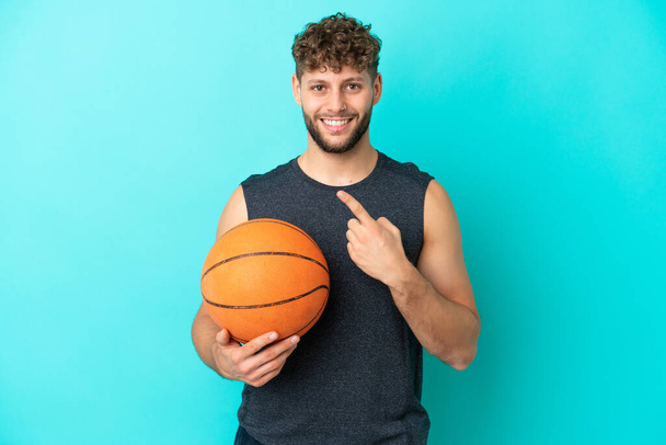 Jóképű fiatalember játszik kosárlabda elszigetelt kék háttér mutatva az oldalon, hogy bemutassa a termék - Fotó, kép
