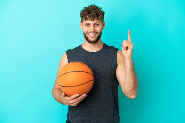 Hezký mladý muž hraje basketbal izolované na modrém pozadí ukazuje a zvedá prst ve znamení toho nejlepšího - Fotografie, Obrázek