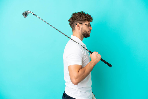 Young handsome caucasian man isolated on blue background playing golf in lateral position - Foto, Imagen