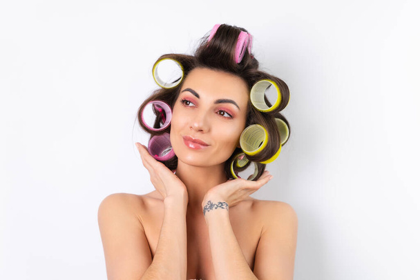 Beautiful housewife. Young cheerful woman with hair curlers and bare shoulders getting ready for a date night. Makes a hairstyle at home on a white background. - Photo, image