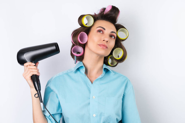 Beautiful housewife. Young cheerful woman with hair curlers and hair dryer getting ready for a date night. Makes a hairstyle at home on a white background. - Фото, изображение