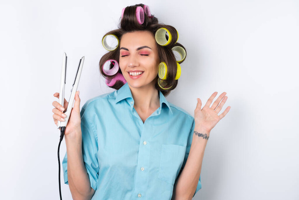 Beautiful housewife. Young cheerful woman with hair curlers and flat iron getting ready for a date night. Makes a hairstyle at home on a white background - 写真・画像