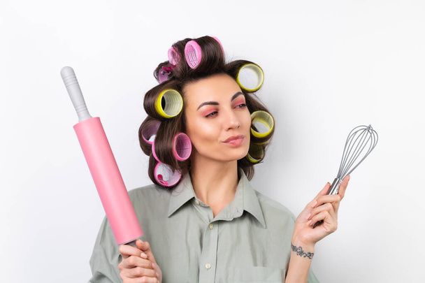 Beautiful housewife. Young cheerful woman with hair curlers, bright make-up, a rolling pin and a whisk in her hands, on a white background. Thinking about what to cook for dinner. - Photo, image
