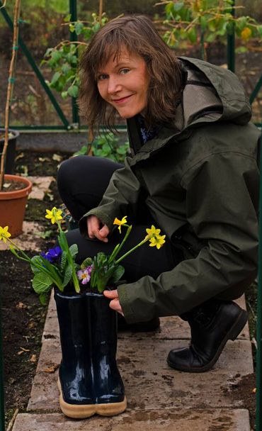 Montrer une femme brune attrayante et souriante plantant des bulbes de printemps dans un planteur de bottes wellingotn. - Photo, image