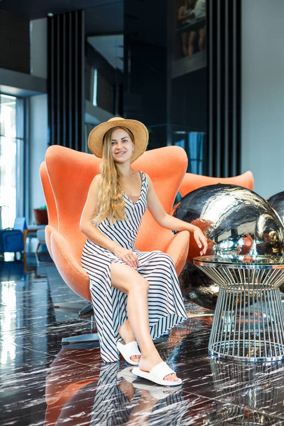 A beautiful young woman in a dress and a hat is sitting in the lobby of a luxury hotel. She is on vacation. - Fotografie, Obrázek
