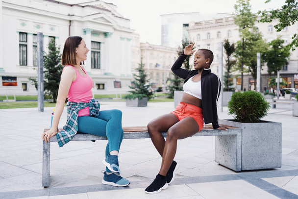 Two multiethnic curvy girls relaxing after exercise session, outdoor communicating. - Photo, Image