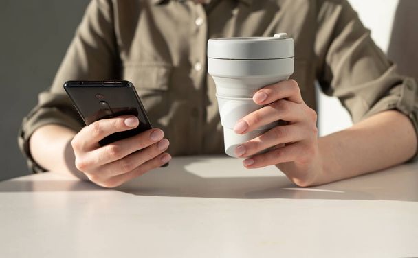 Woman hands holding phone and eco cup. Closeup. Woman sitting at table and using smartphone for typing message, surfing Internet, paying online - Photo, Image