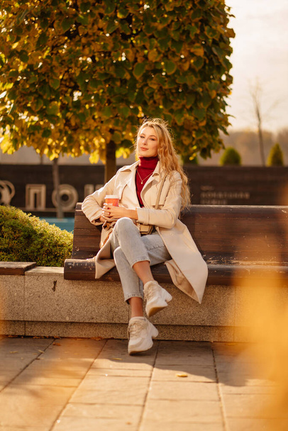 a beautiful blonde with a cup of coffee sits on a bench in an autumn park. - Foto, afbeelding