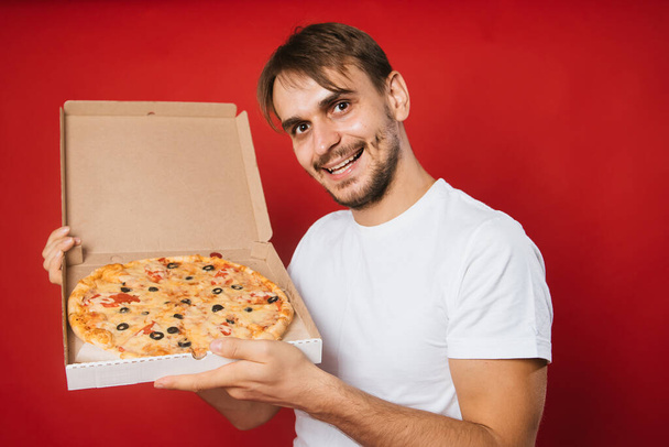A man with a smile in a white t-shirt on a red background holds a box with an appetizing pizza in his hands. Pizza delivery man. Pizza deliveryman. Mock-up - Zdjęcie, obraz