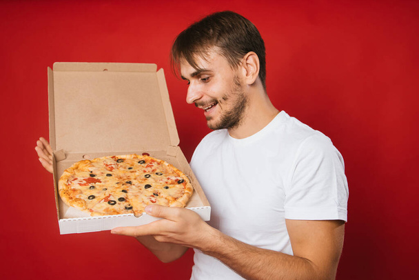 A man with a smile in a white t-shirt on a red background holds a box with an appetizing pizza in his hands. Pizza delivery man. Pizza deliveryman. Mock-up - Photo, Image