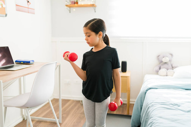 Beautiful young girl working out with dumbbells weights. Cute kid with a healthy lifestyle exercising in her bedroom - Foto, imagen