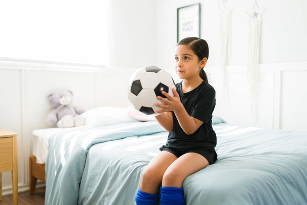 Adorable little child holding a ball while sitting on her bed and ready to go to a football soccer practice  - Photo, Image