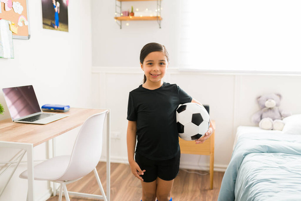 Excited young girl smiling at her bedroom while thinking of becoming a soccer player when she grows up  - Foto, immagini