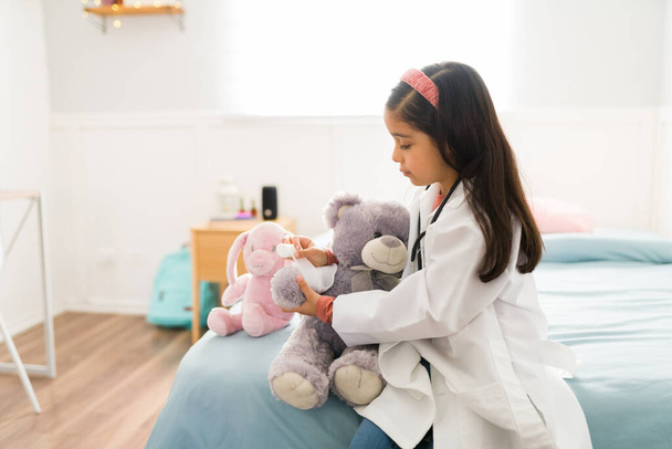 Urocza młoda dziewczyna gra lekarza w sypialni i nakłada bandaż na pluszowego misia  - Zdjęcie, obraz