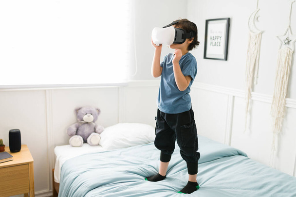 Διασκεδάζω. Παιχνιδιάρικο παιδί που στέκεται στο κρεβάτι του και χρησιμοποιεί ακουστικά VR και παρακολουθεί ένα βιντεοπαιχνίδι εικονικής πραγματικότητας  - Φωτογραφία, εικόνα