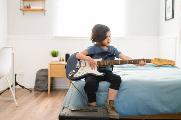 Schattige jonge jongen zitten op zijn bed tijdens het spelen van de elektrische gitaar en het leren van een nieuw lied - Foto, afbeelding