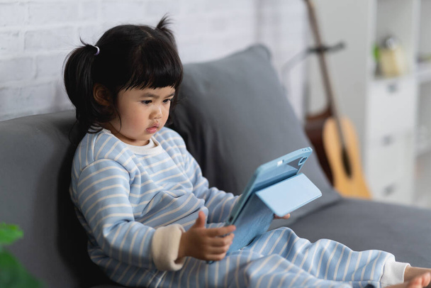 Aasian vauva tyttö näyttää ja kosketa tabletti istuu sohvalla. Pikkutyttö pelaa peliä käyttäen pöydän kotona. Teknologia perhe vauva käsite. - Valokuva, kuva