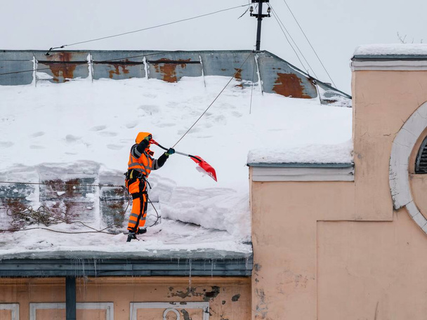 Ένας εργάτης με λαμπερή φόρμα με ζώνη ασφαλείας με φτυάρι καθαρίζει το χιόνι από την οροφή ενός παλιού κτιρίου.. - Φωτογραφία, εικόνα
