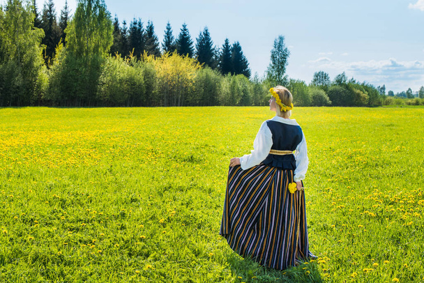 Young woman in national clothes wearing yellow dandelion wreath in spring field. Springtime - Valokuva, kuva