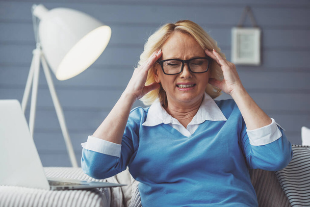 Beautiful tired old woman in casual clothes and glasses is having a headache while working with laptop at home - Φωτογραφία, εικόνα