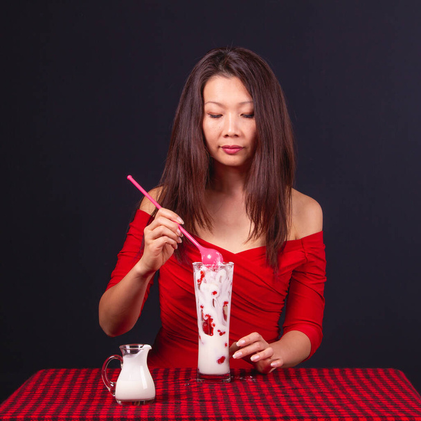 Piękna Azjatka w średnim wieku siedzi przy stole z obrusem w kratkę i je pyszny jogurt z truskawkami.. - Zdjęcie, obraz