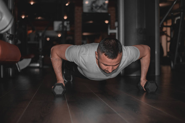 Man doing workout with heavy dumbbell. Confident muscled young man wearing sport wear and doing plank position while exercising on the floor in loft interior. - Photo, Image