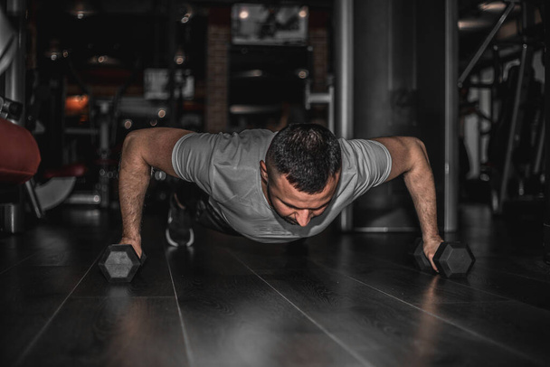 Az ember nehéz súlyzóval edz. Magabiztos izmos fiatalember sport viselet és csinál deszka pozíciót, miközben gyakorol a padlón padlástérben. - Fotó, kép