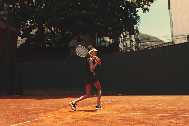 A sportswoman holding a tennis racket over her shoulder, with her feet flexed and looking straight ahead. An African woman practicing tennis attacks alone outdoors. The lady exercising on a game court - 写真・画像