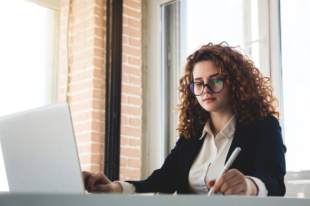 Portret of a successful young curly red-haired woman in a business suit and glasses works in an office on a laptop - Fotografie, Obrázek