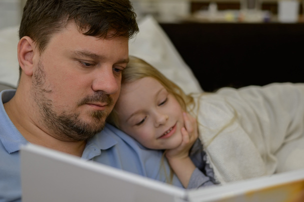 papá entretiene al niño durante la enfermedad: mira, lee un libro, un libro de fotos con su hija en casa. El padre sospecha que su hija está enferma. El niño no se siente bien. el concepto de - Foto, imagen