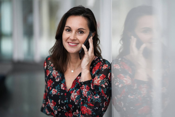 happy smiling chic business lady in red floral dress making mobile phone call and looking straight into the camera with a modern office background - Фото, зображення